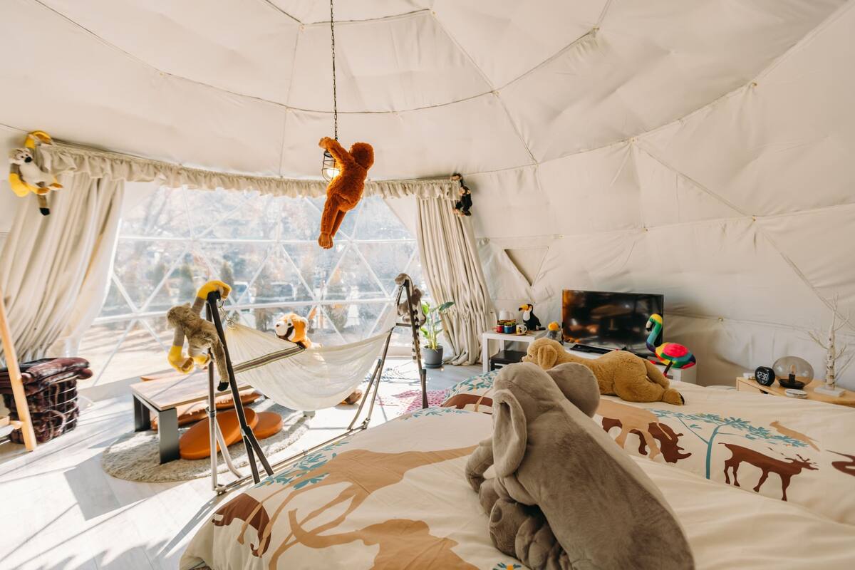 A cozy, well-lit tent-style room with a bed, hanging hammock, and various stuffed animals.