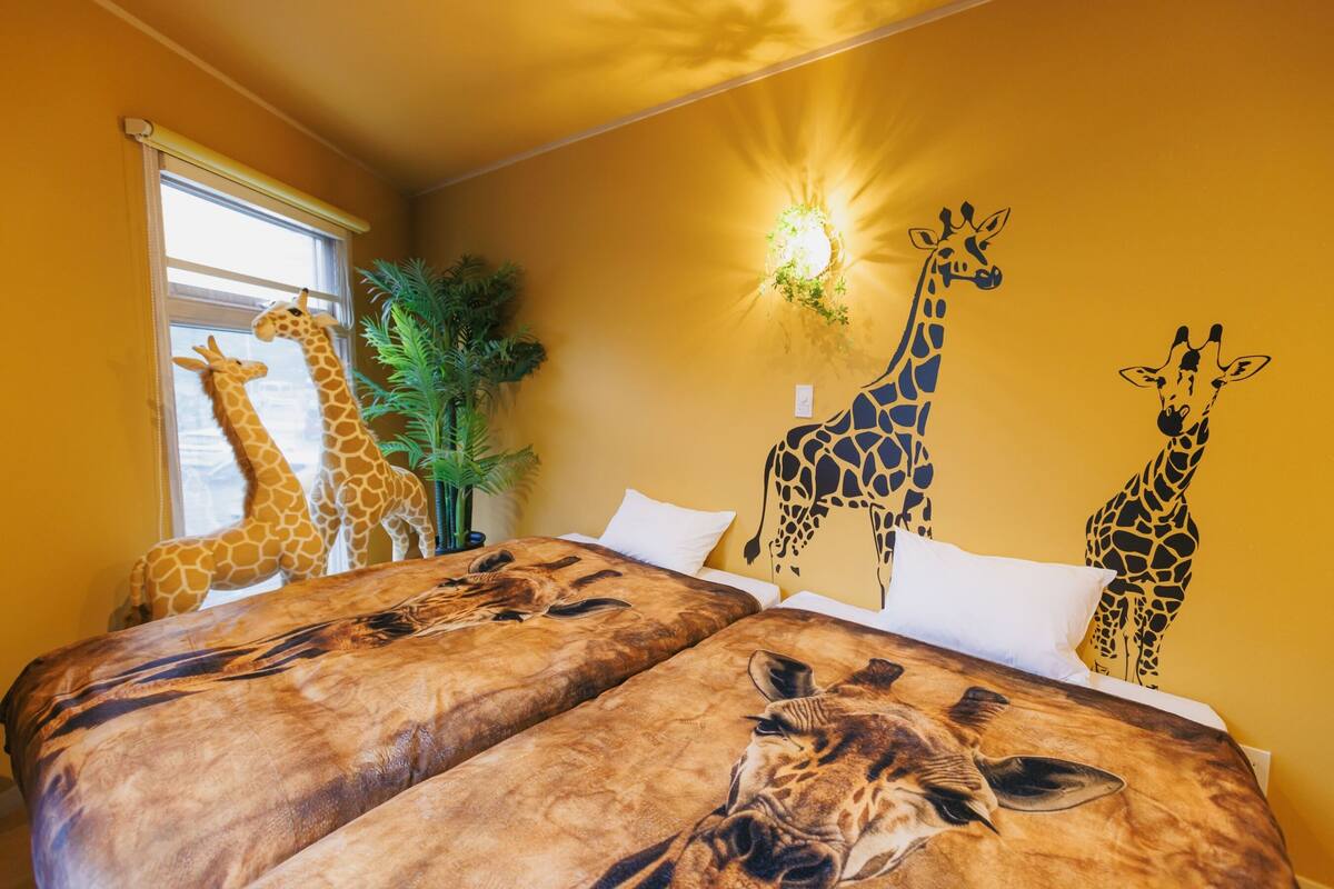A room with a bed featuring giraffe-themed bedding, a wall-mounted light fixture, and a window with a view.