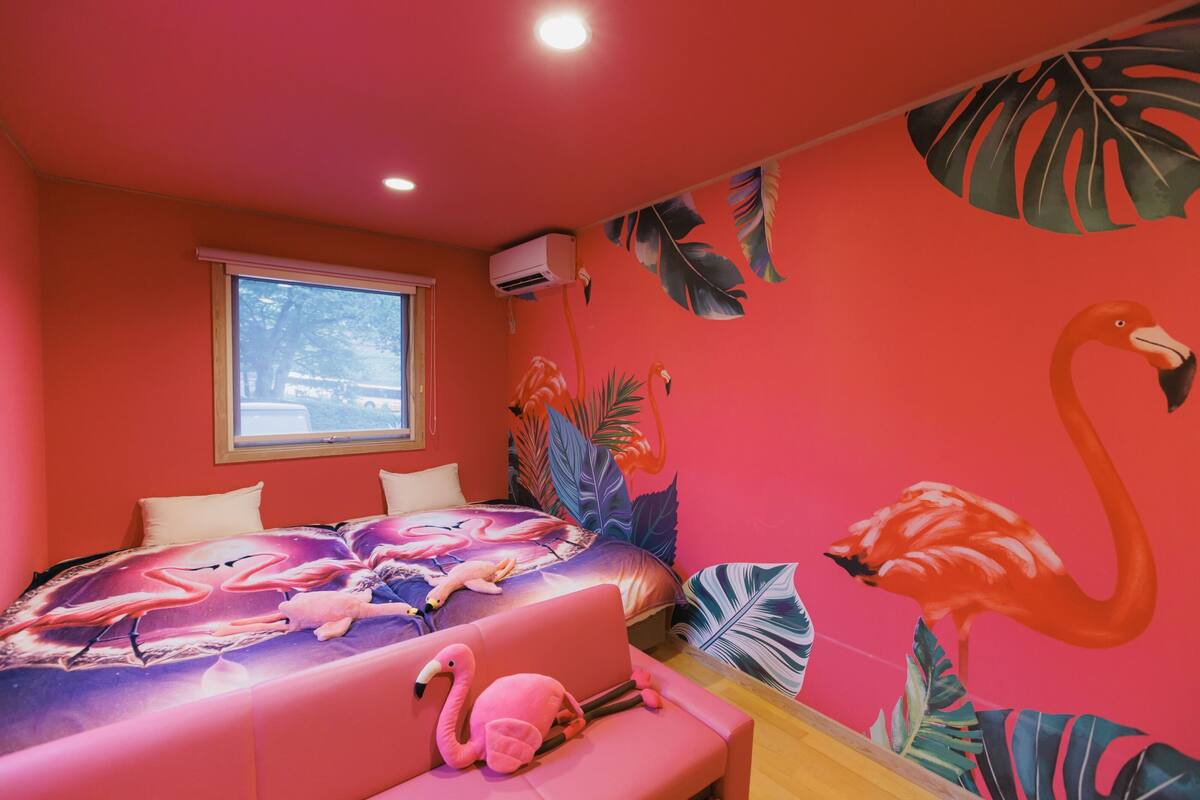 A room with a bed, a window, an air conditioning unit, and a wall mural of flamingos and tropical leaves.