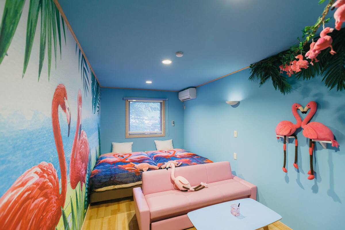 A room with a bed, a pink sofa, a small table, a window, and a wall mural.