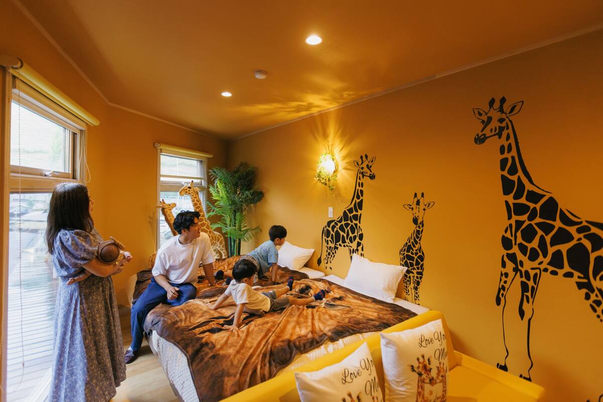 A room with a bed, giraffe wall art, a window with blinds, and a plant.