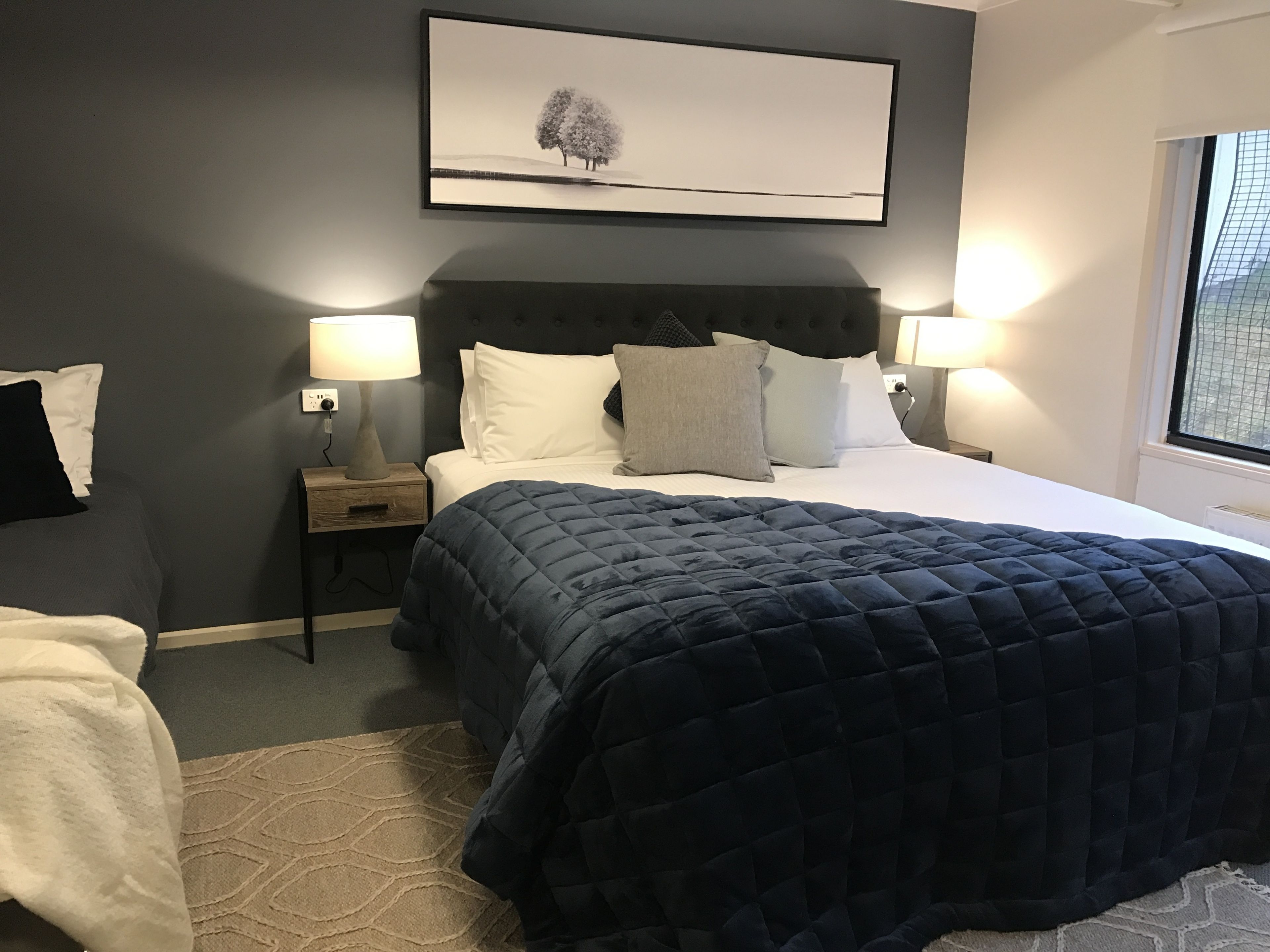 A neatly made bed with a dark quilt, two bedside lamps, a framed picture on the wall, and a window with a view.