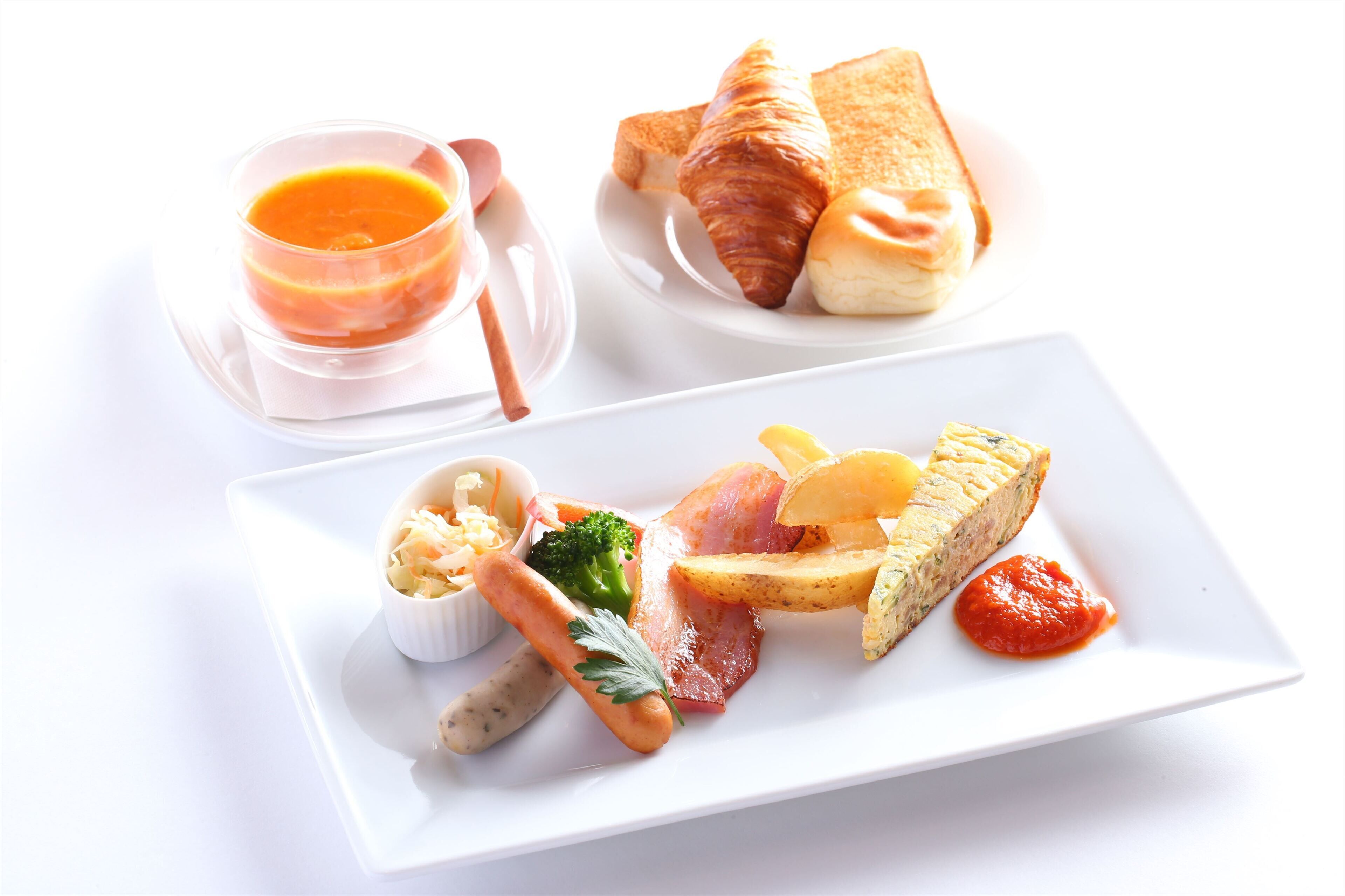 A white plate with a variety of food items including a croissant, a piece of bread, a small bowl of soup, a sausage, a piece of meat, a carrot, and a tomato sauce.