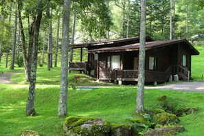 A rustic cabin with a porch, surrounded by tall trees and lush greenery. - Tateshina Tokyu Hotel (Chino)