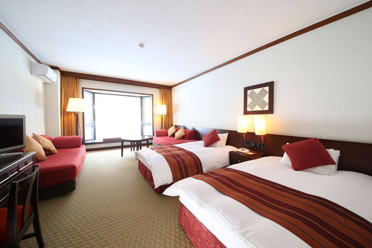 Una habitación de hotel con dos camas, televisor, escritorio y ventana con cortinas.