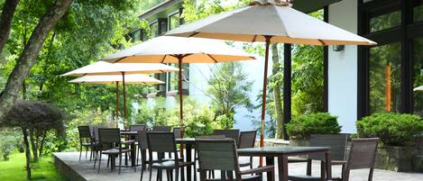 An outdoor patio with tables and chairs, surrounded by greenery and a building with large windows.