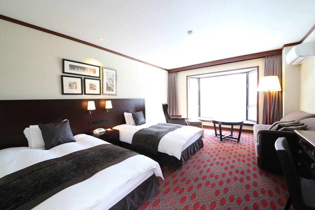 Habitación de hotel con dos camas, un escritorio, una silla, un ventanal grande y una alfombra roja con diseño.