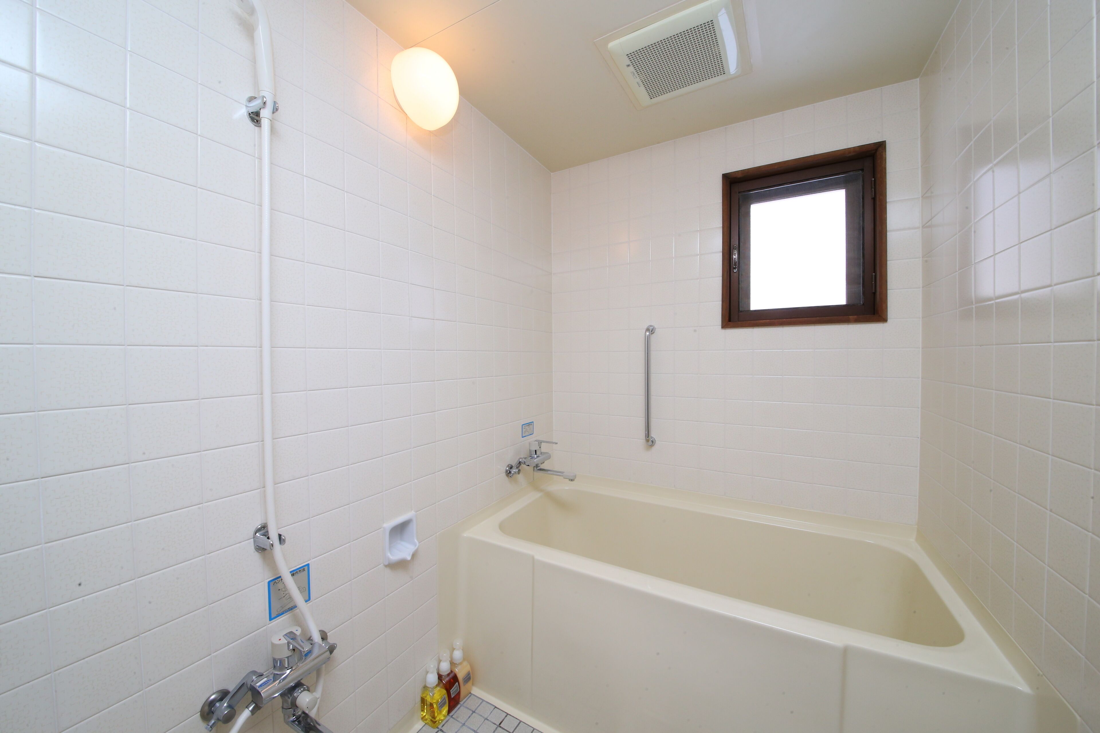 A bathroom with white tiled walls, a bathtub, a showerhead, a window, and a handrail.