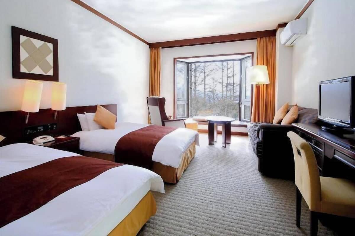 A hotel room with two beds, a desk with a television, a chair, a lamp, and a view of the outdoors.