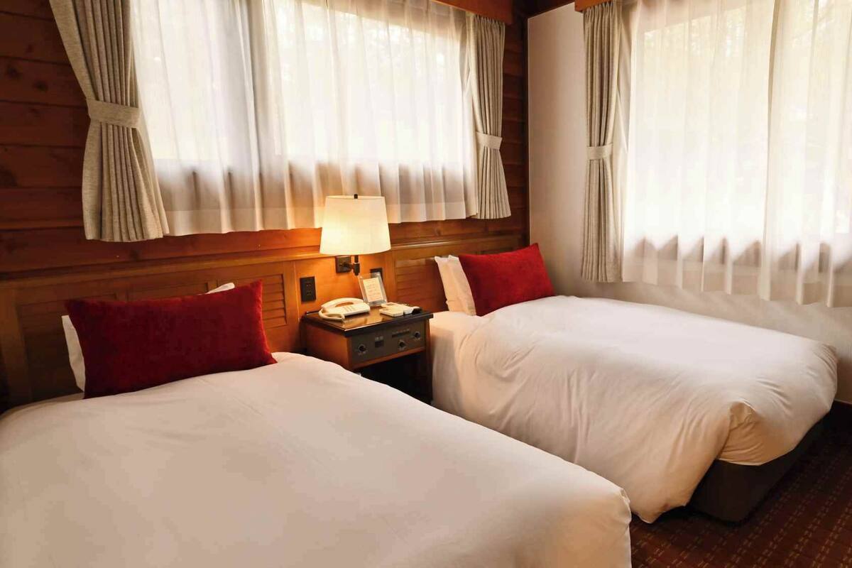 Una habitación de hotel con dos camas, una mesita de noche con lámpara y cortinas en las ventanas.