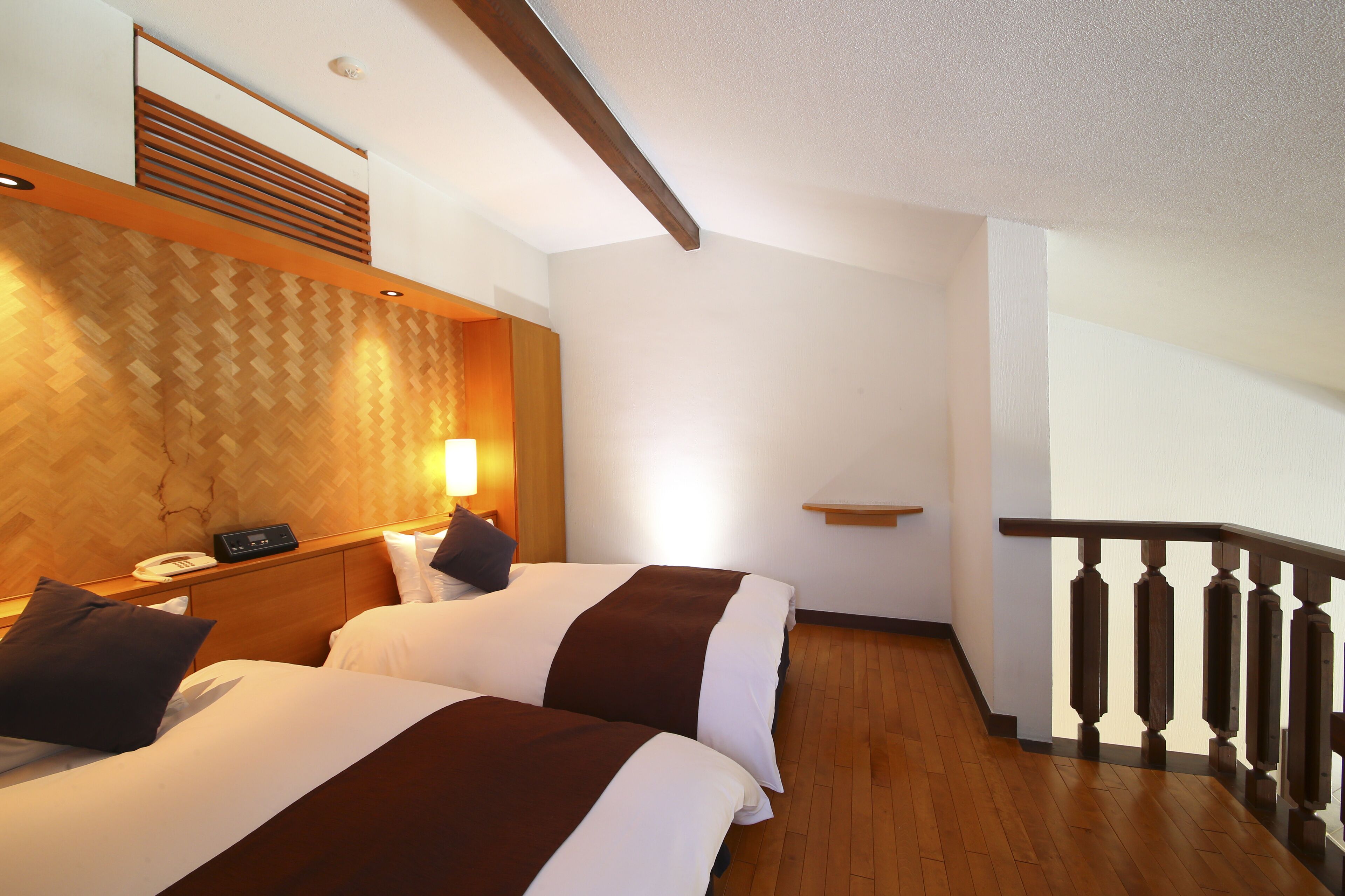 A hotel room with two beds, wooden flooring, a wooden headboard, a bedside table with a lamp, and a wooden railing on the balcony.