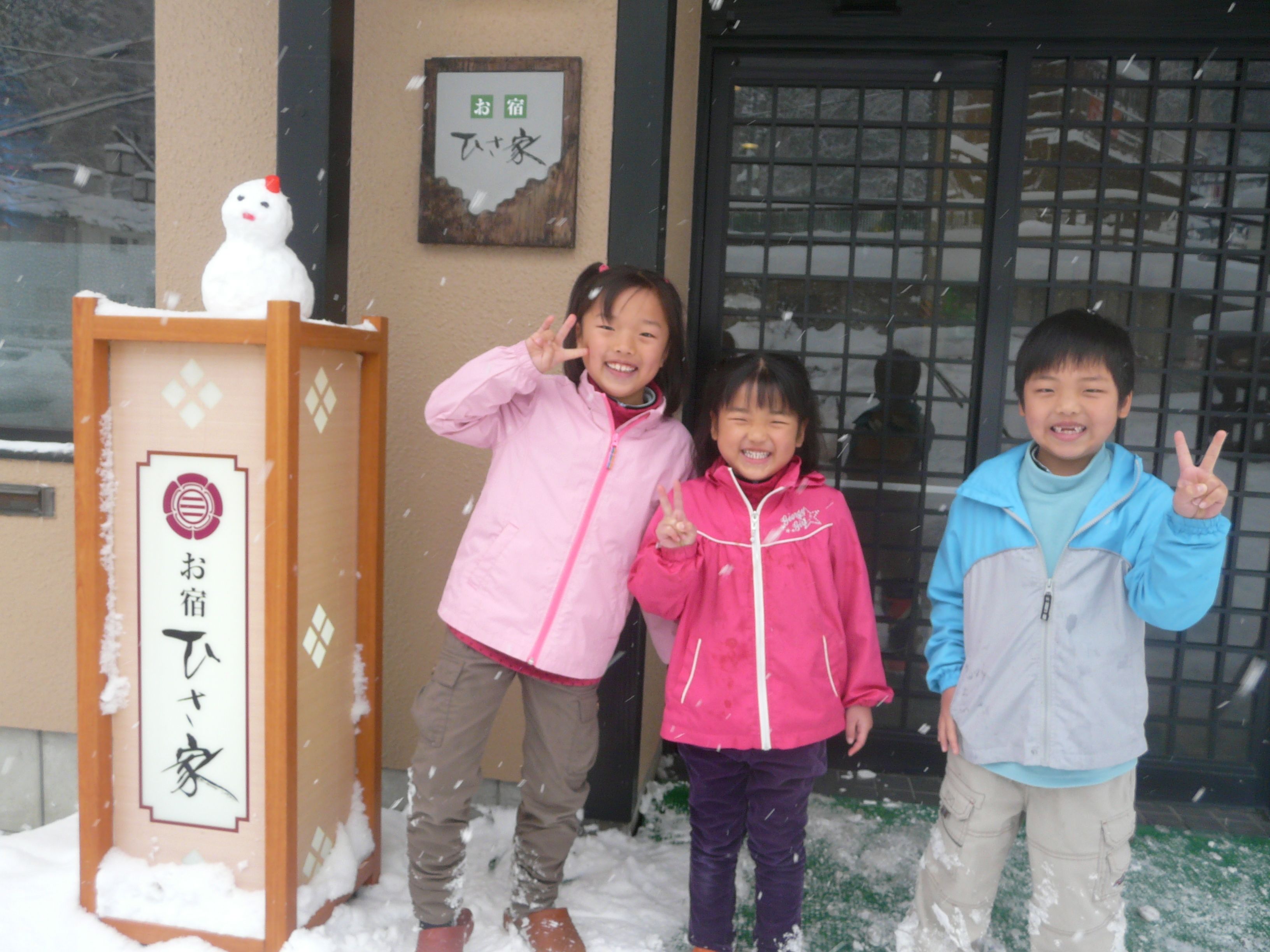 Three children standing in front of a building with a wooden sign and a black gate.