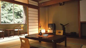 Una habitación japonesa tradicional con suelo de tatami, una mesa baja, sillas, un televisor y una ventana con cortinas.