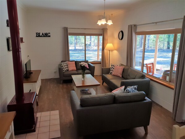 A modern living room with two sofas, a coffee table, and a lamp. There is a large window with a view of trees and a clock on the wall. - Woodlane Cottages (Nulkaba)
