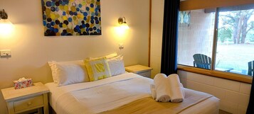 Una cama bien hecha con sábanas blancas y una toalla doblada, una mesita de noche con un dispensador de pañuelos, un cuadro en la pared y una ventana con vistas al exterior.