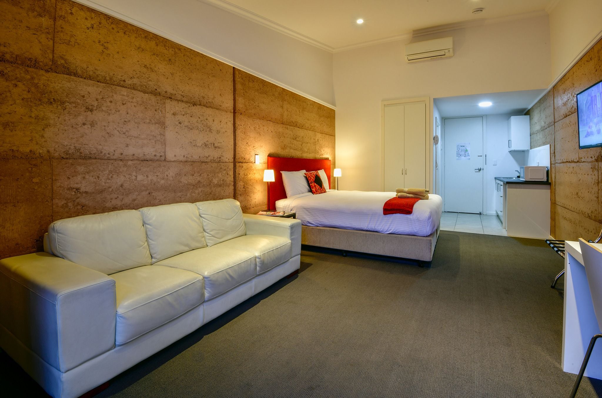 A hotel room with a white sofa, a bed with red bedding, and a kitchenette area.