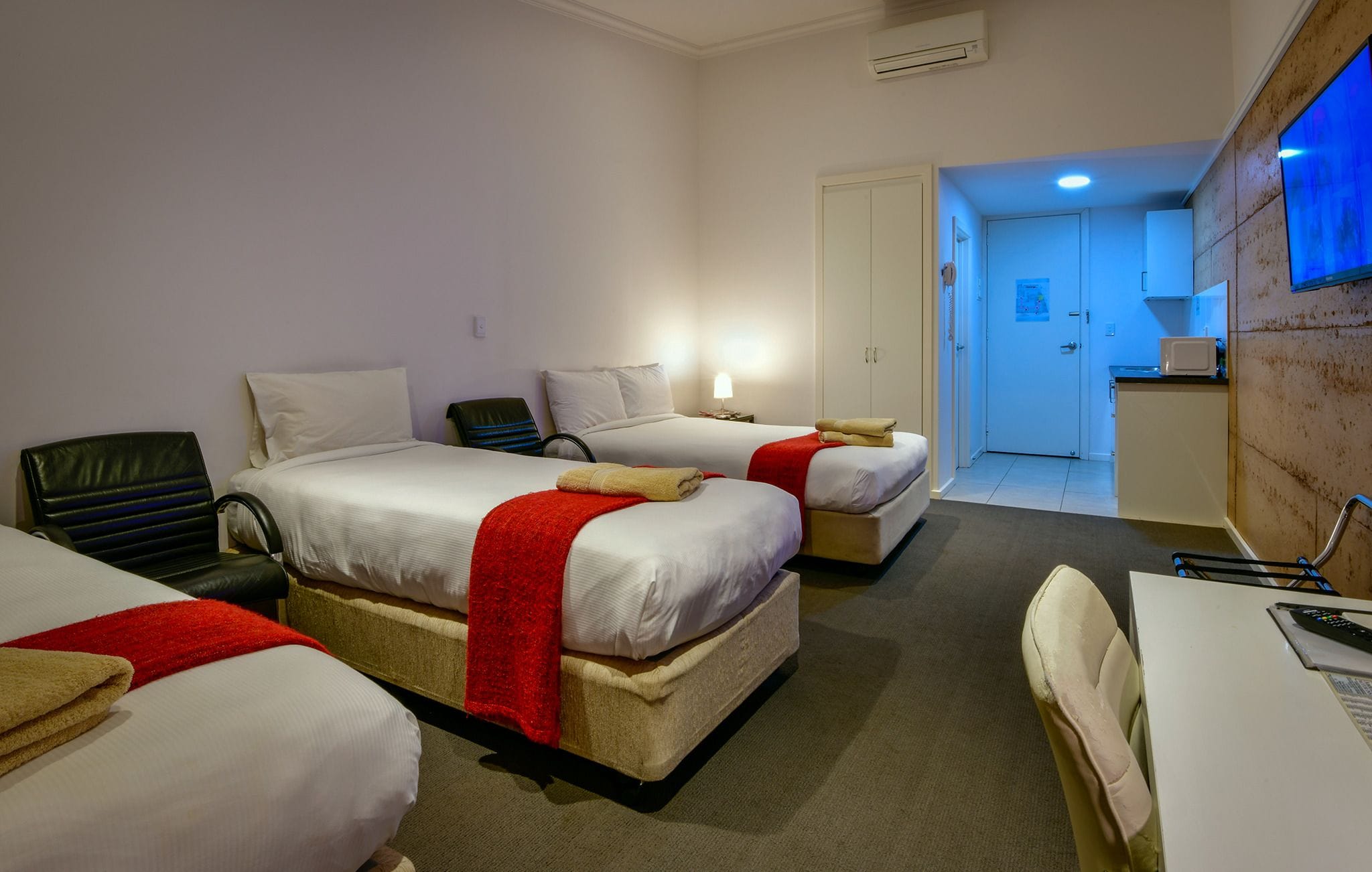 A hotel room with two beds, a desk, a chair, a small kitchenette, and a bathroom visible through an open door.