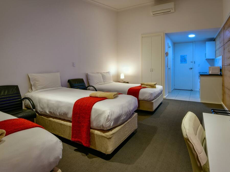 A hotel room with two beds, a desk, a chair, a small kitchenette, and a bathroom visible through an open door.