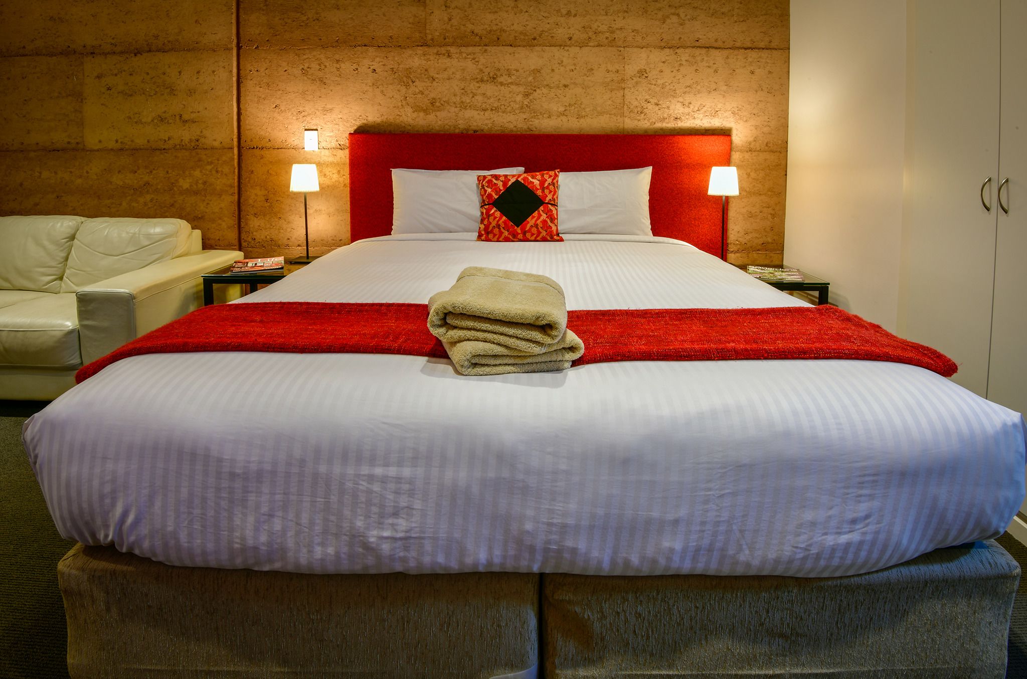 Une chambre d’hôtel avec un grand lit, deux lampes de chevet, une tête de lit rouge et un plaid beige sur le lit.