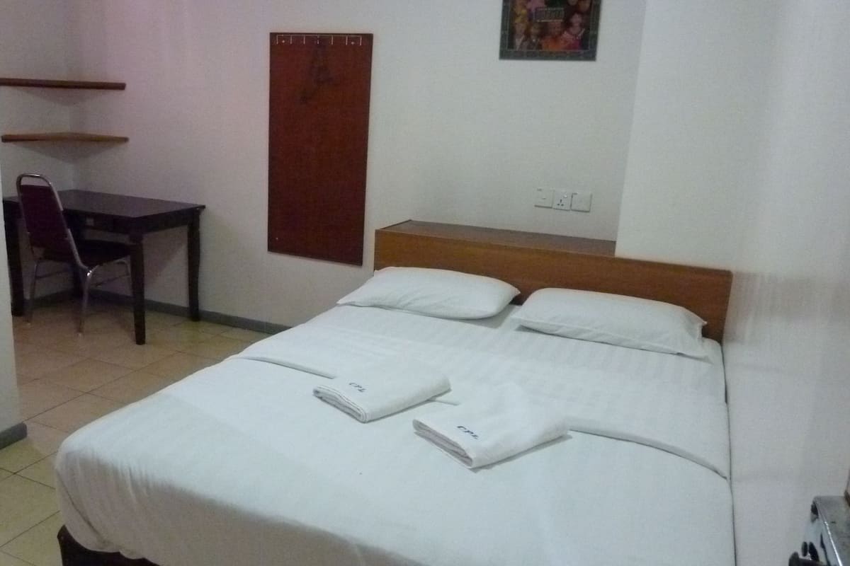A neatly made bed with white linens and two folded towels, a wooden headboard, a small desk with a chair, and a wall-mounted shelf.