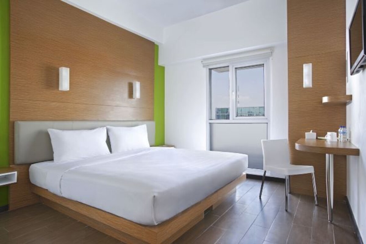 Quarto de hotel moderno com cabeceira de madeira, cama grande, uma mesa pequena com uma cadeira e janela com vista para a cidade.