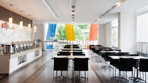 A modern cafeteria with long tables and chairs, a buffet counter with food containers, and large windows with a view of greenery.