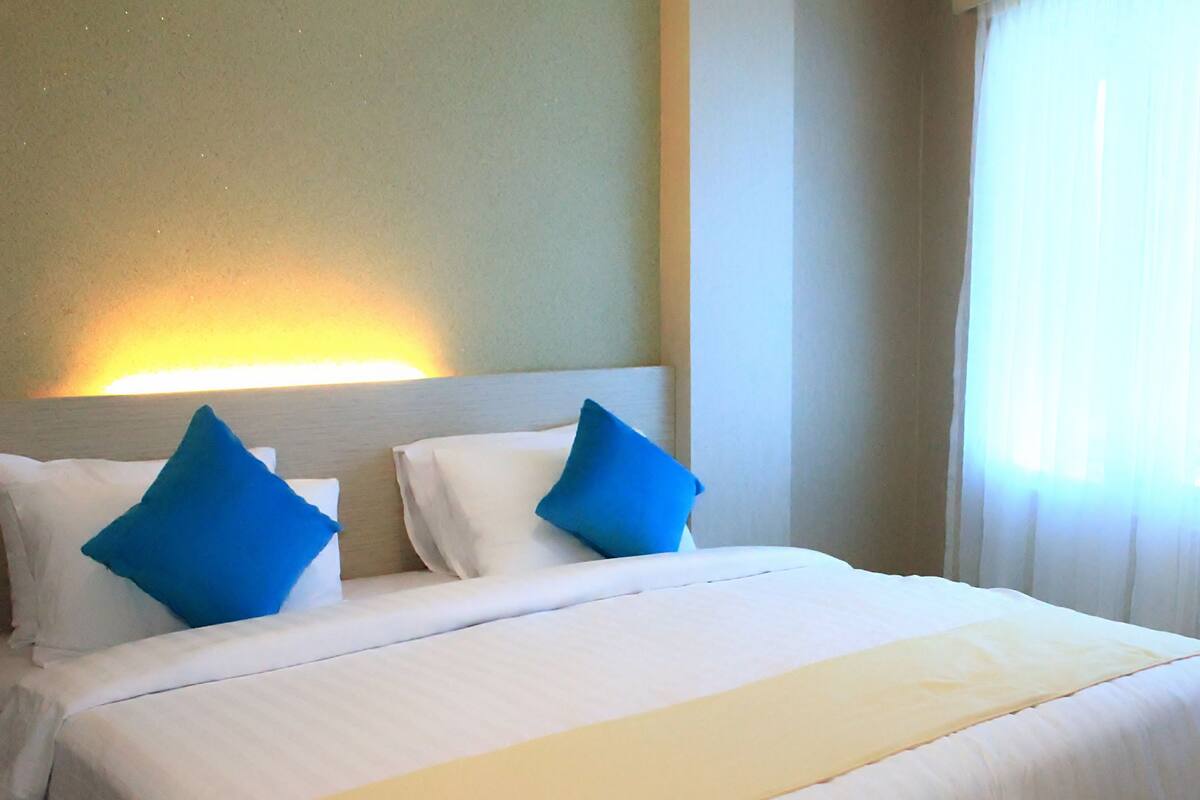 Une chambre d’hôtel avec un lit bien fait, une tête de lit et une fenêtre ornée de rideaux légers.