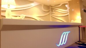 Un espace d’accueil au design moderne, comprenant un comptoir de réception blanc orné d’un logo bleu rétroéclairé, une lampe décorative et un mur aux motifs abstraits.