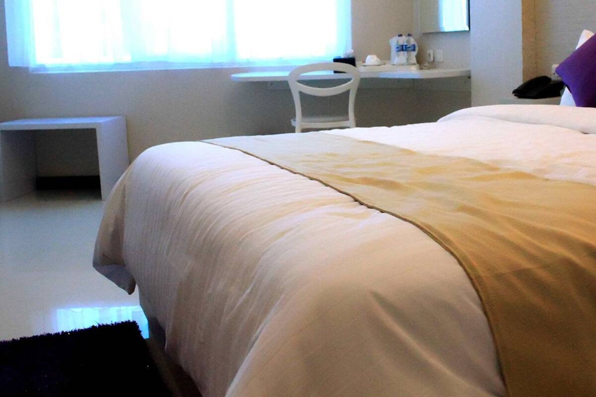 Un lit bien fait, recouvert d’une couvre-lit beige et blanche, une petite table de chevet et une chaise dans une chambre d’hôtel.
