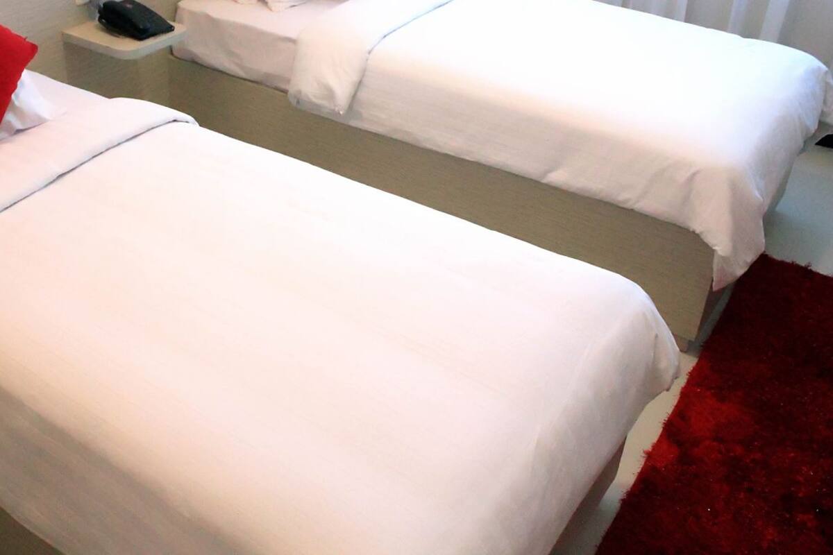 Deux lits simples avec du linge de lit blanc et des oreillers rouges, un tapis rouge et une petite table de chevet avec une tél écommande.