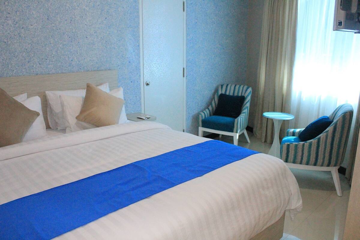 Une chambre d’hôtel avec un grand lit, deux fauteuils, une petite table et un téléviseur.