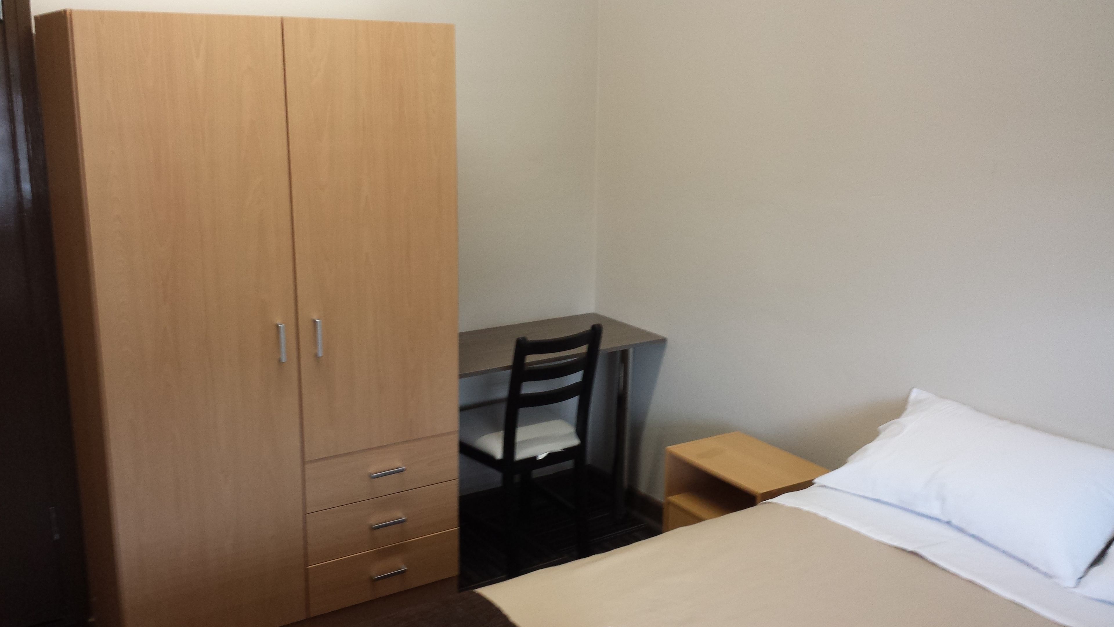 A bedroom with a bed, a wooden wardrobe, a desk, and a chair.