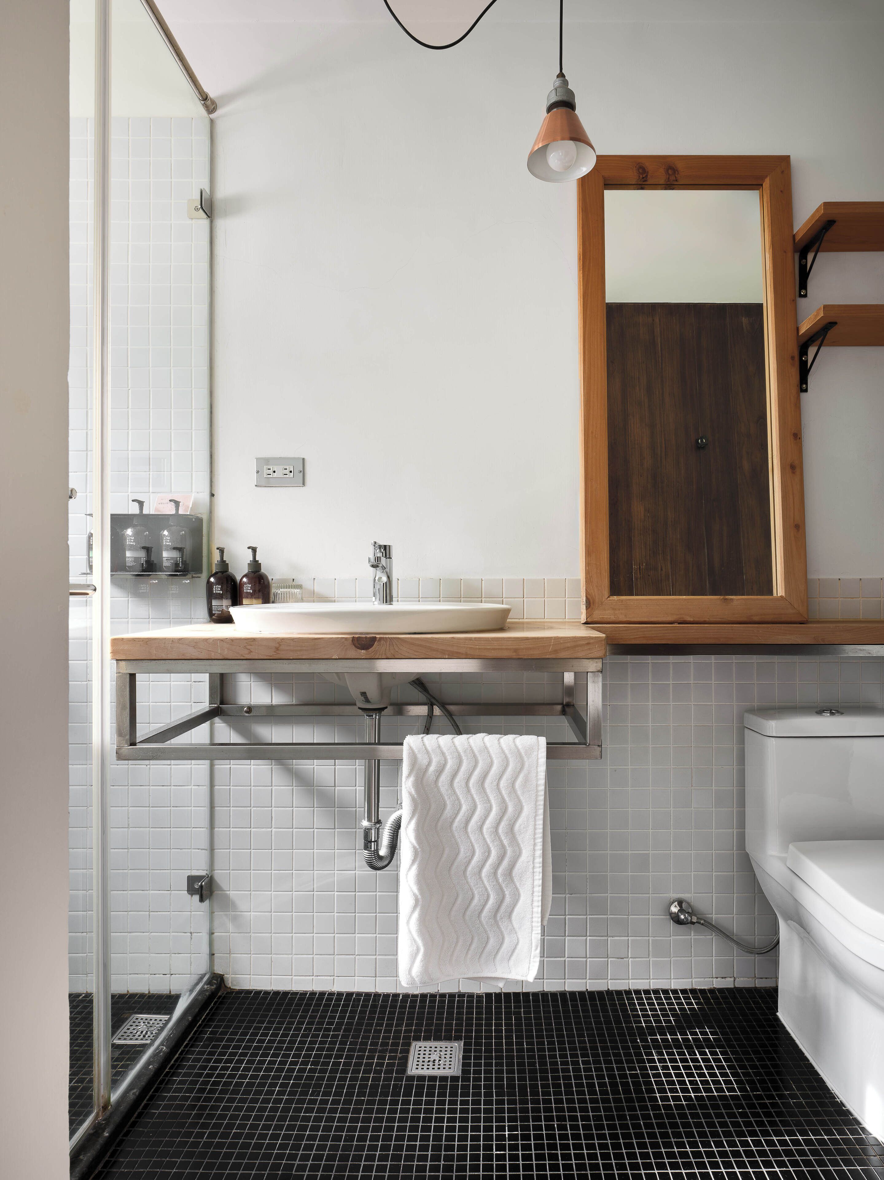 A bathroom with a white sink, wooden countertop, and a mirror above it. There is a towel hanging on a rack, and a shower with a black tiled floor.
