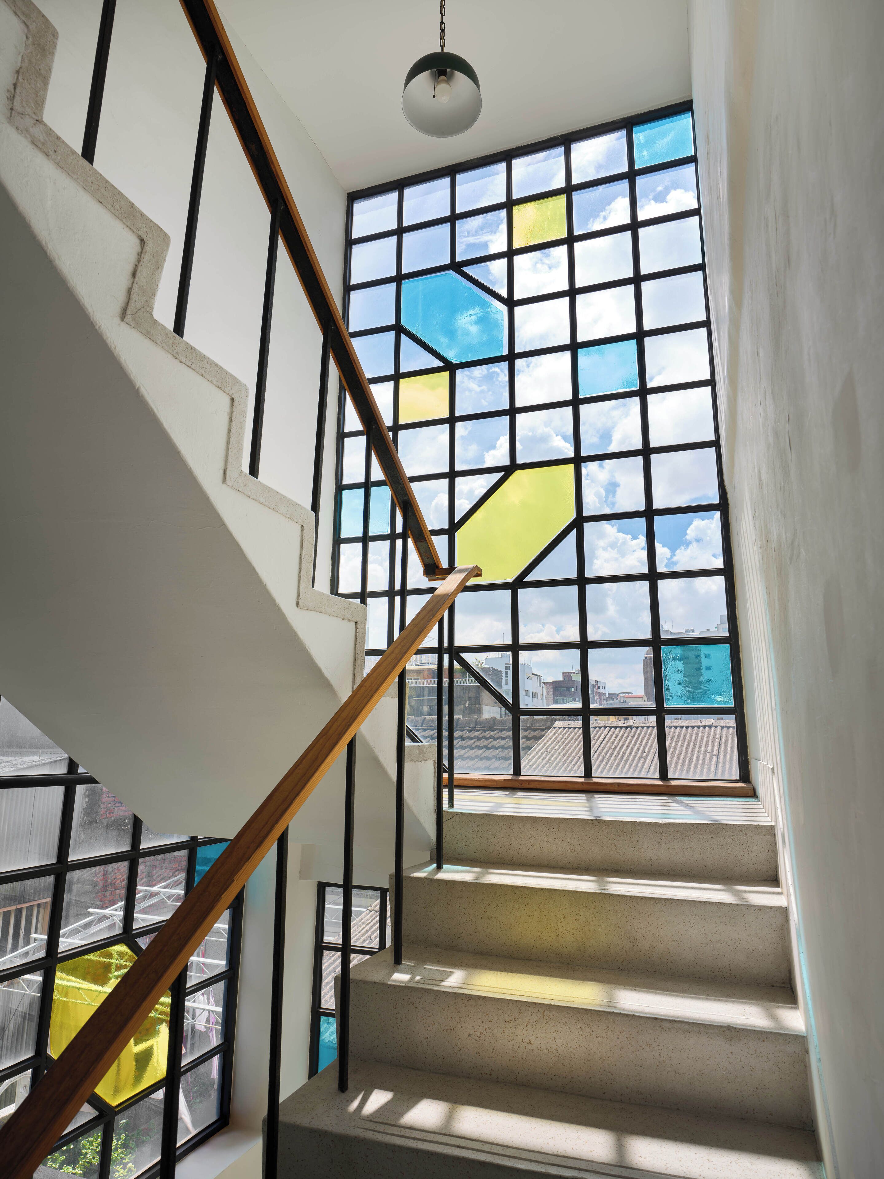 A staircase with a geometric stained glass window, a wooden handrail, and a hanging light fixture.
