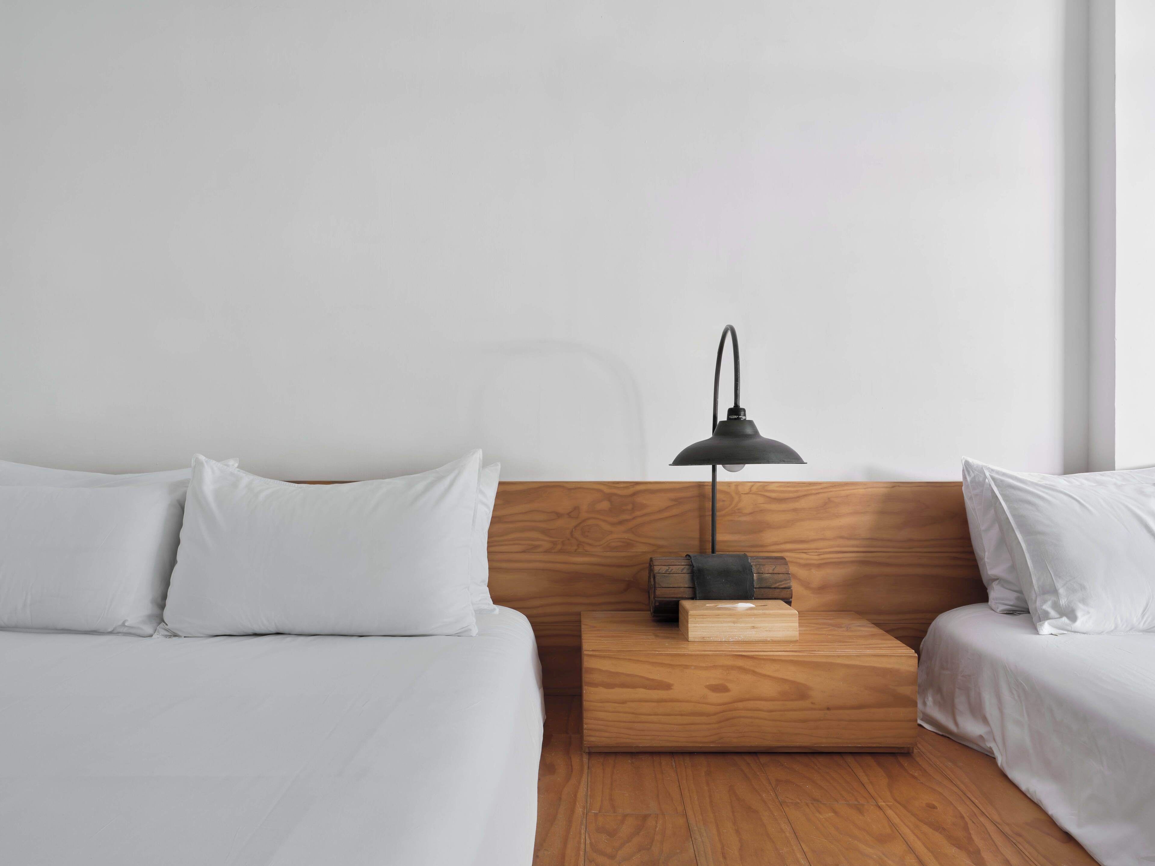 A minimalist hotel room with a wooden headboard, white bedding, and a single bedside table with a lamp.