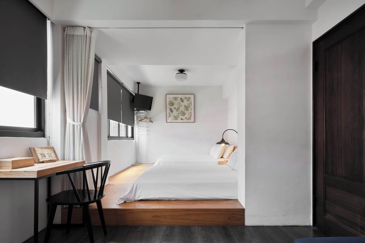 A compact hotel room with a bed, a desk with a chair, a TV, and a window with curtains.