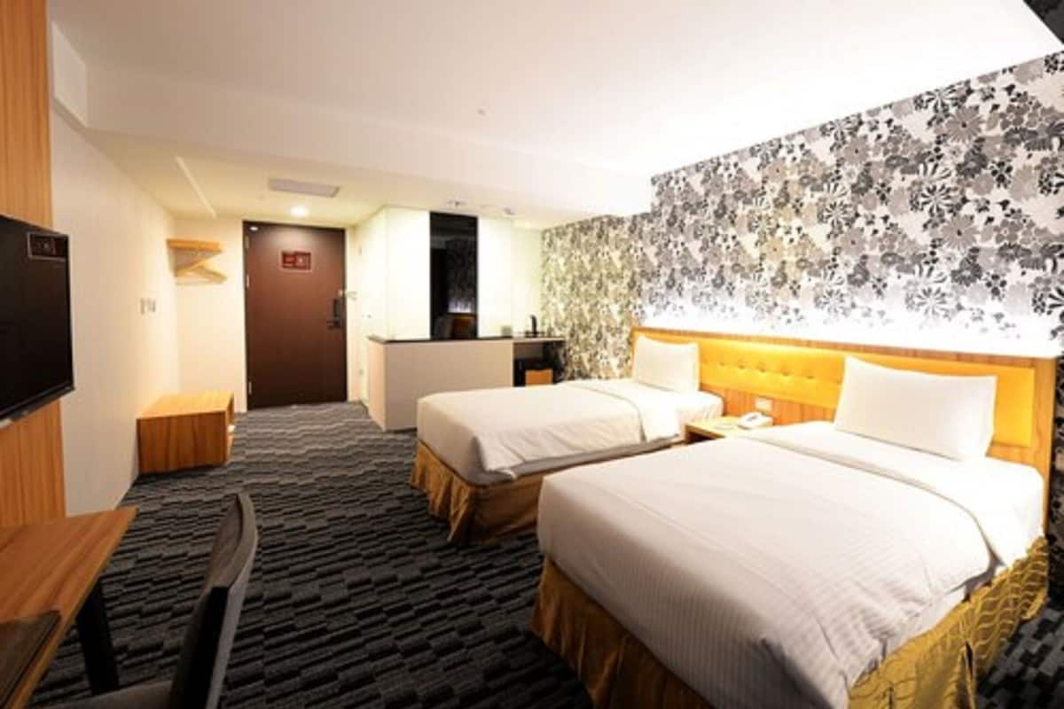Quarto de hotel com duas camas, uma escrivaninha, uma cadeira, uma televisão e parede estampada.