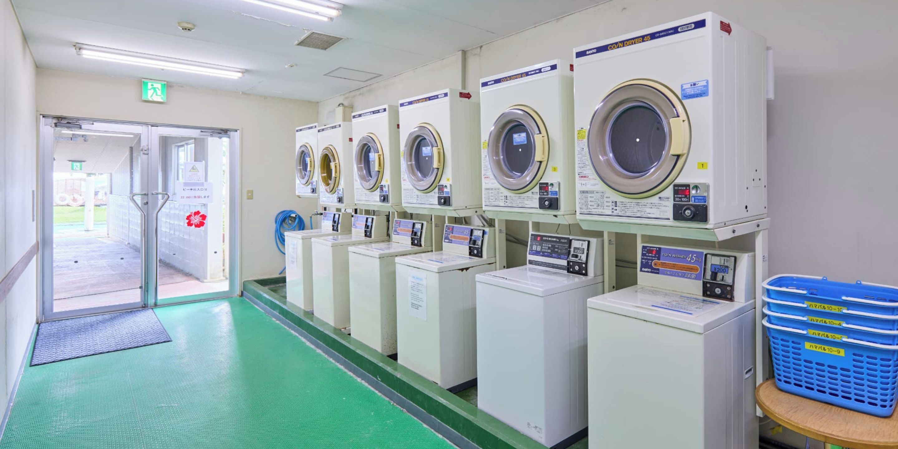 A laundry facility with multiple washing machines and dryers, a green floor, and a door leading outside.