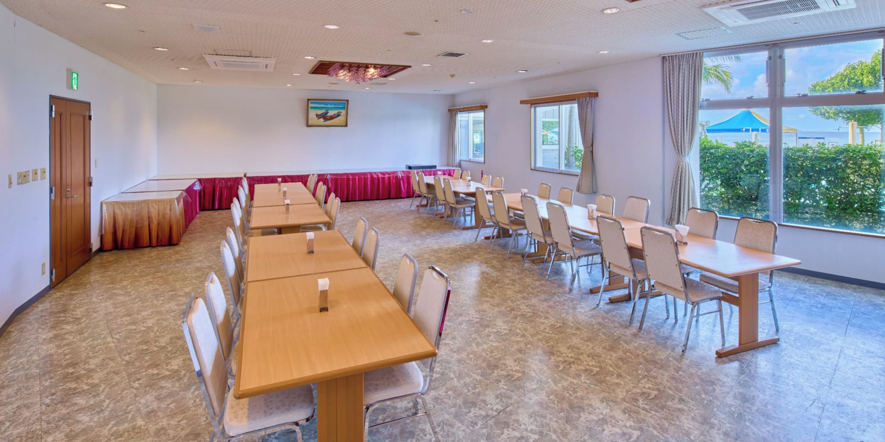 A conference room with multiple tables and chairs, large windows with a view of greenery, and a ceiling with recessed lighting.