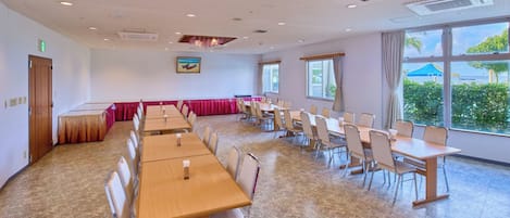 A conference room with multiple tables and chairs, large windows with a view of greenery, and a ceiling with recessed lighting.