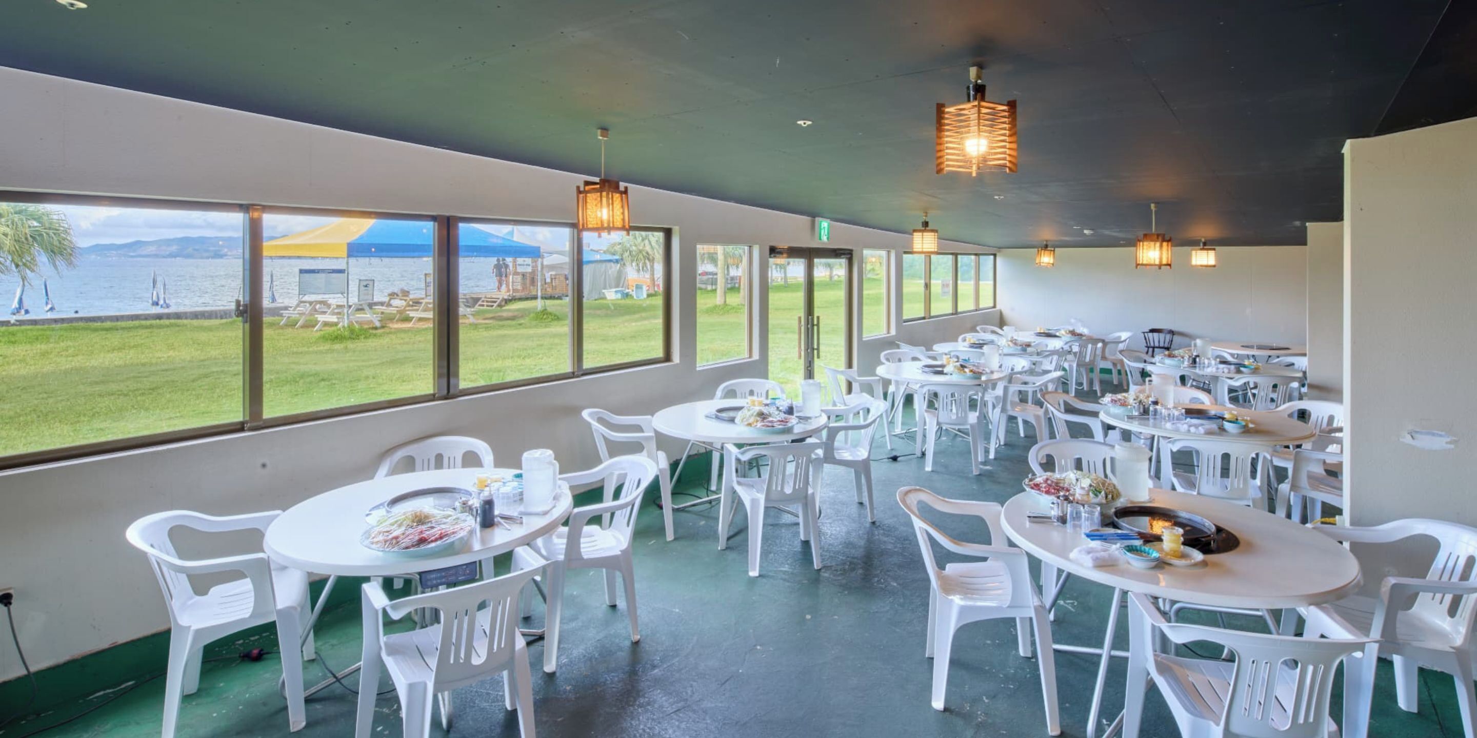 Un restaurant avec des chaises et des tables en plastique blanc, de grandes fenêtres offrant une vue sur la mer et les montagnes, et des lampes suspendues.