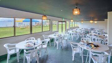 Un restaurant avec des chaises et des tables en plastique blanc, de grandes fenêtres offrant une vue sur la mer et les montagnes, et des lampes suspendues.