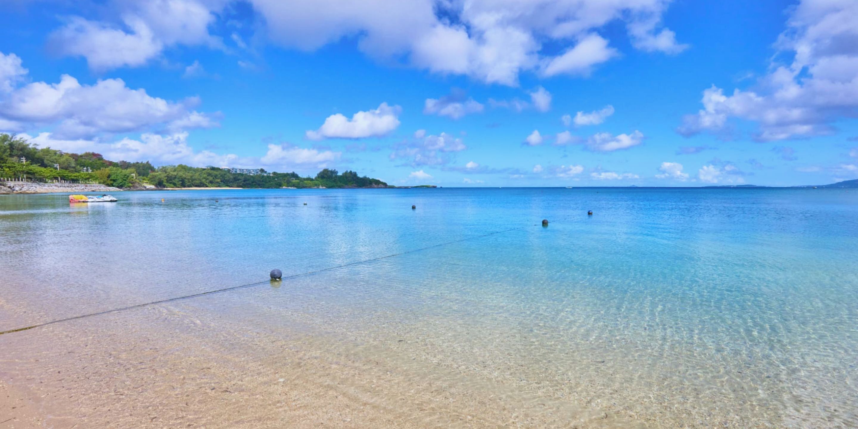 A clear beach with calm waters, a sandy shore, and a distant shoreline with vegetation.