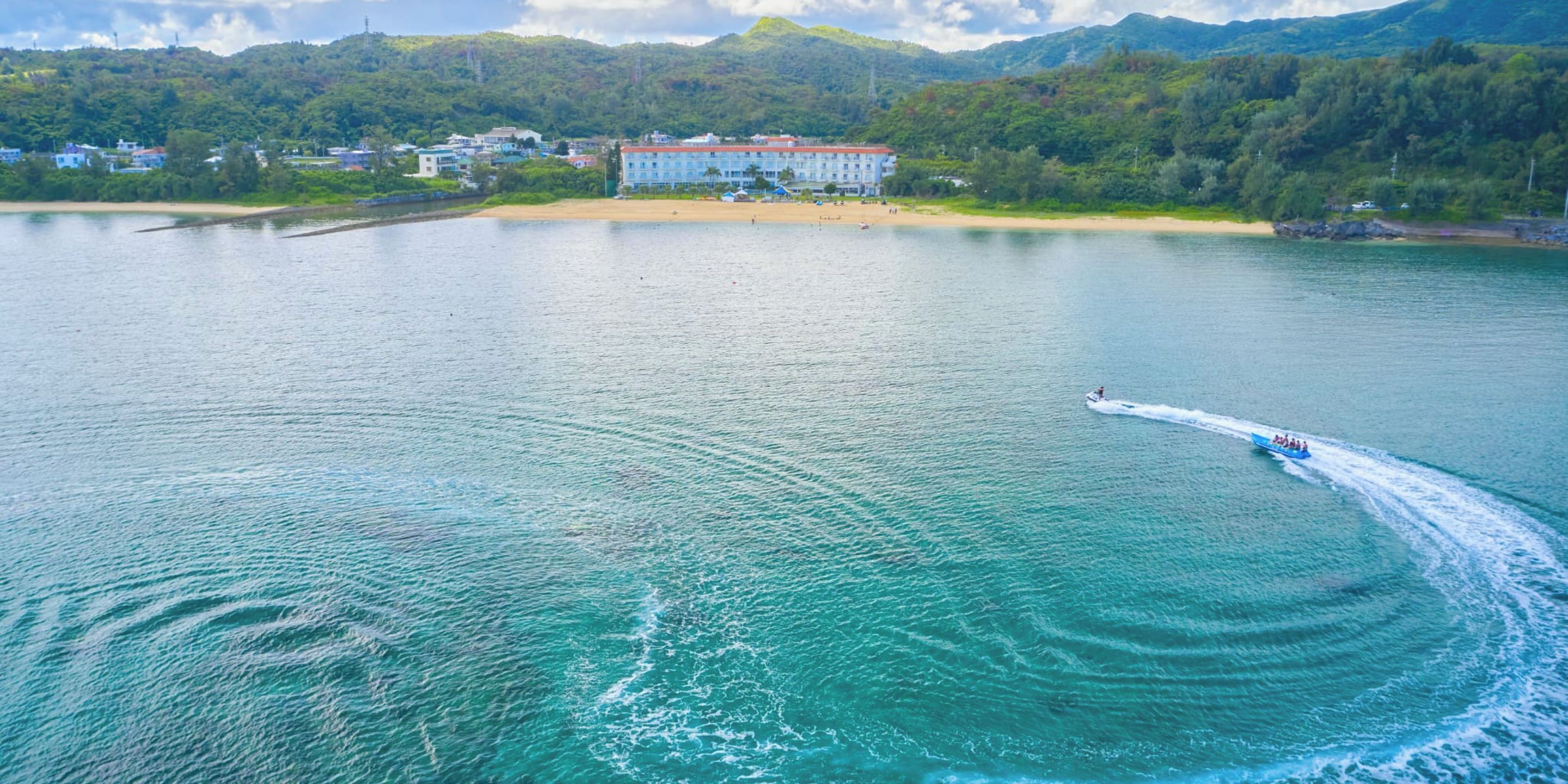 A large body of water with a boat creating a wake. In the background, there is a building with a red roof and a beach with people.
