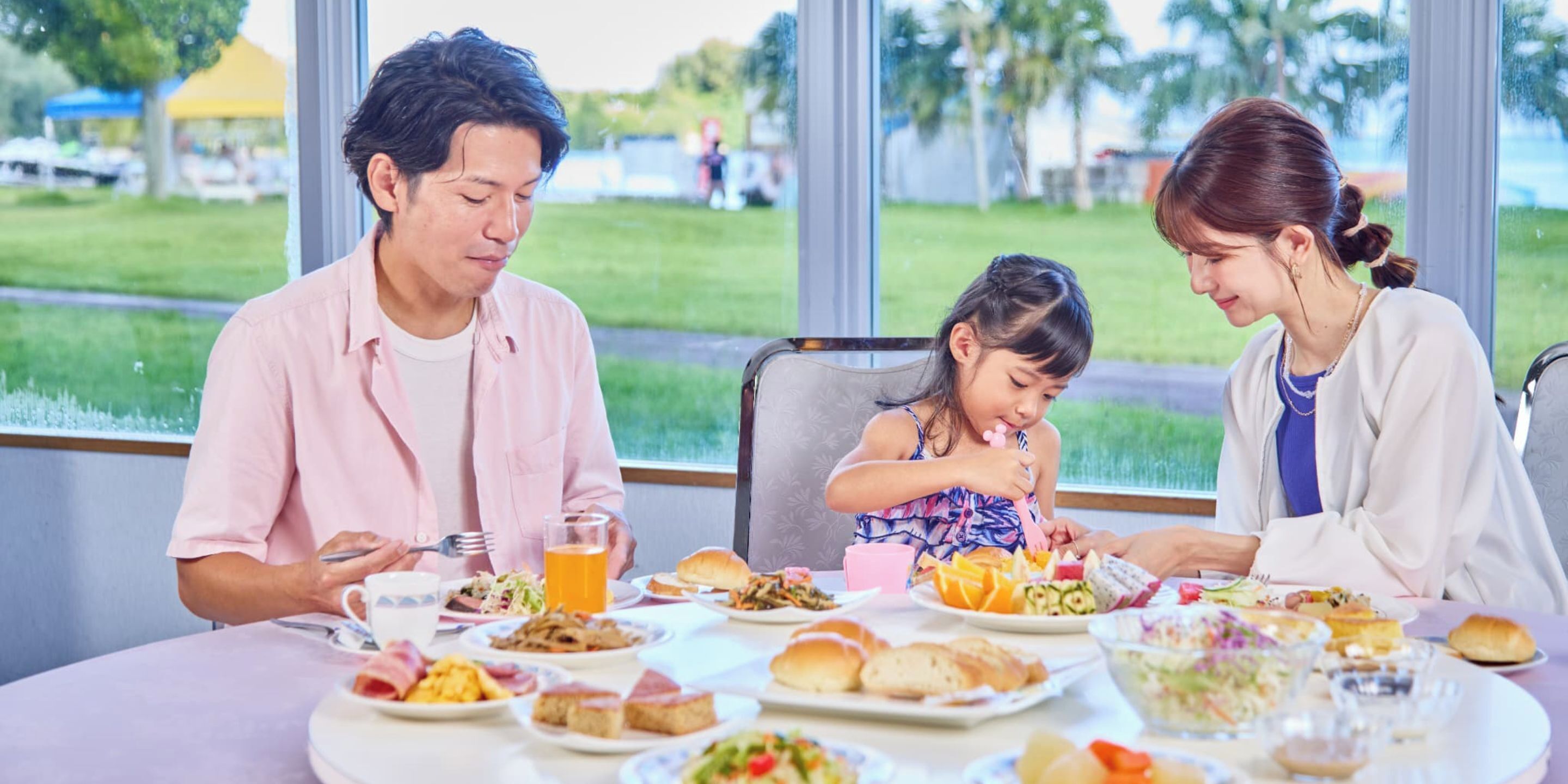 Une famille est assise à une table, devant différents plats, un verre de jus d’orange, et peut admirer un espace verdoyant à l’extérieur.