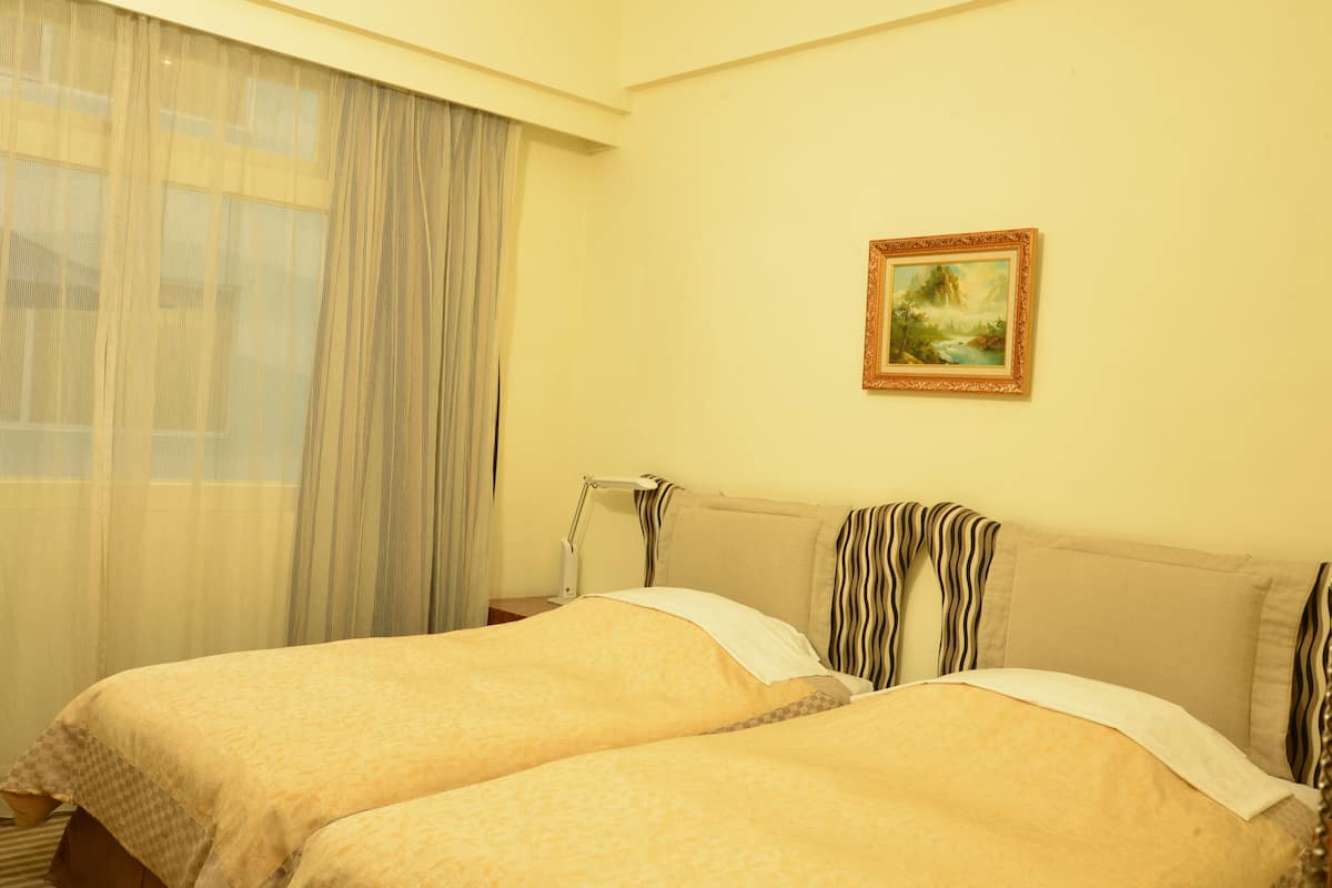 A hotel room with two beds, a painting on the wall, and a window with sheer curtains.