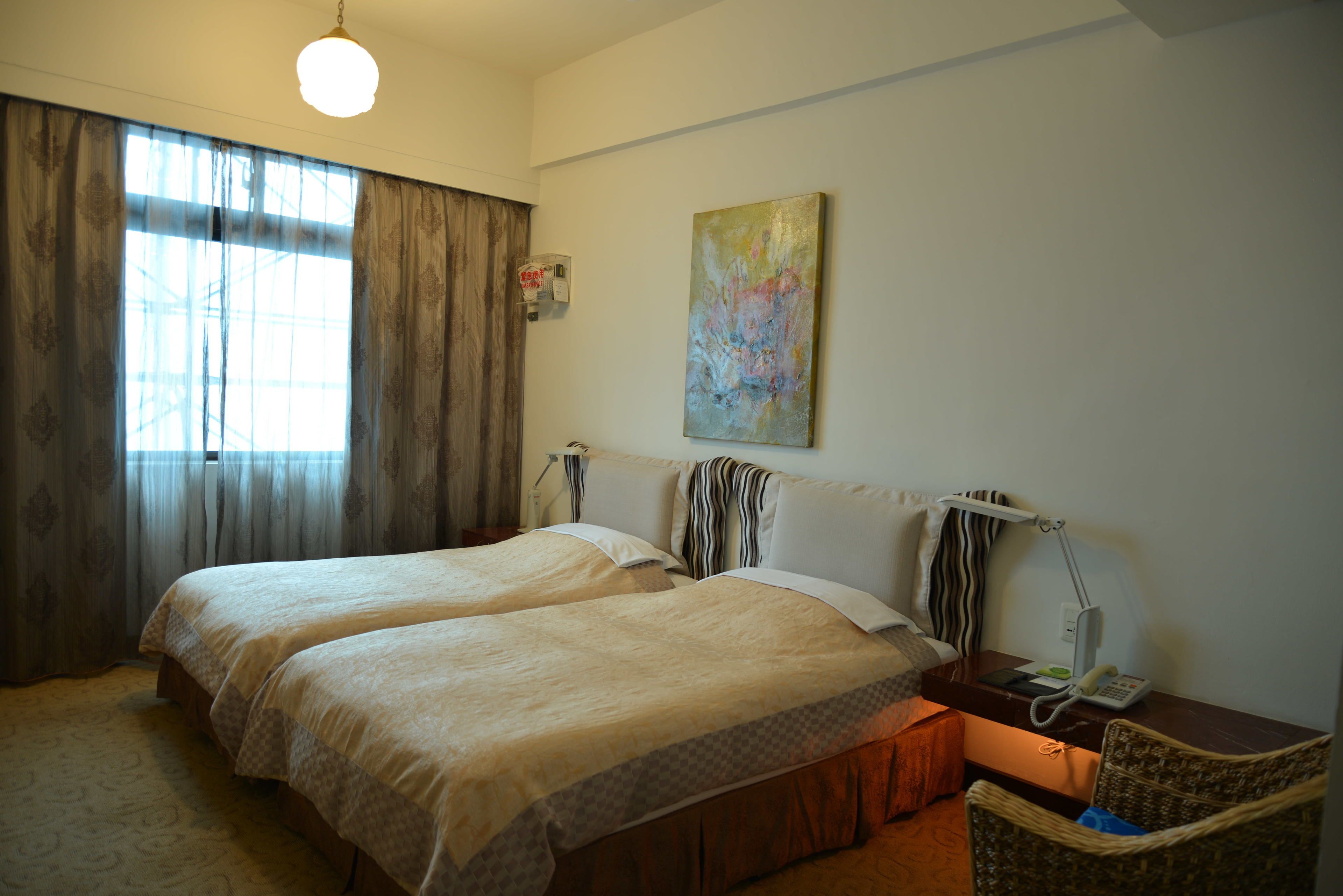 A hotel room with two beds, a bedside table with a lamp, a painting on the wall, and a window with curtains.