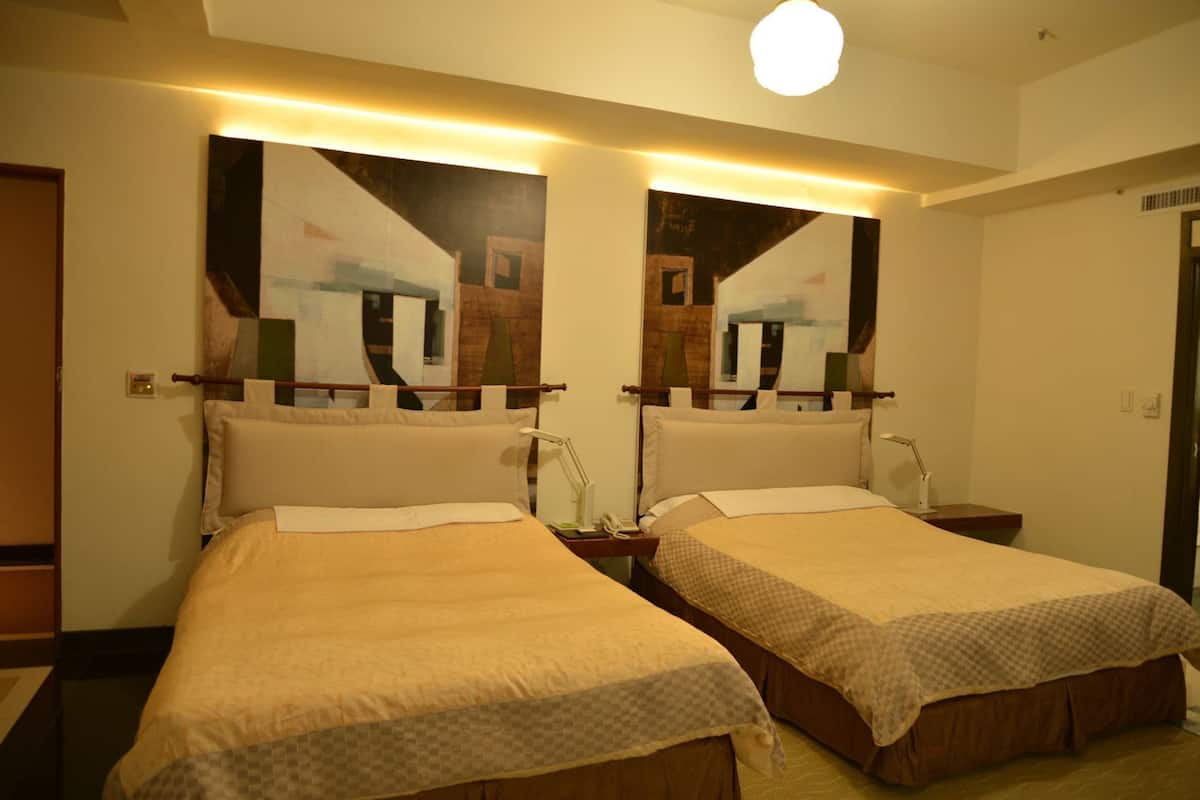 A hotel room with two beds, each with a headboard, bedside tables, lamps, and a wall-mounted light fixture.
