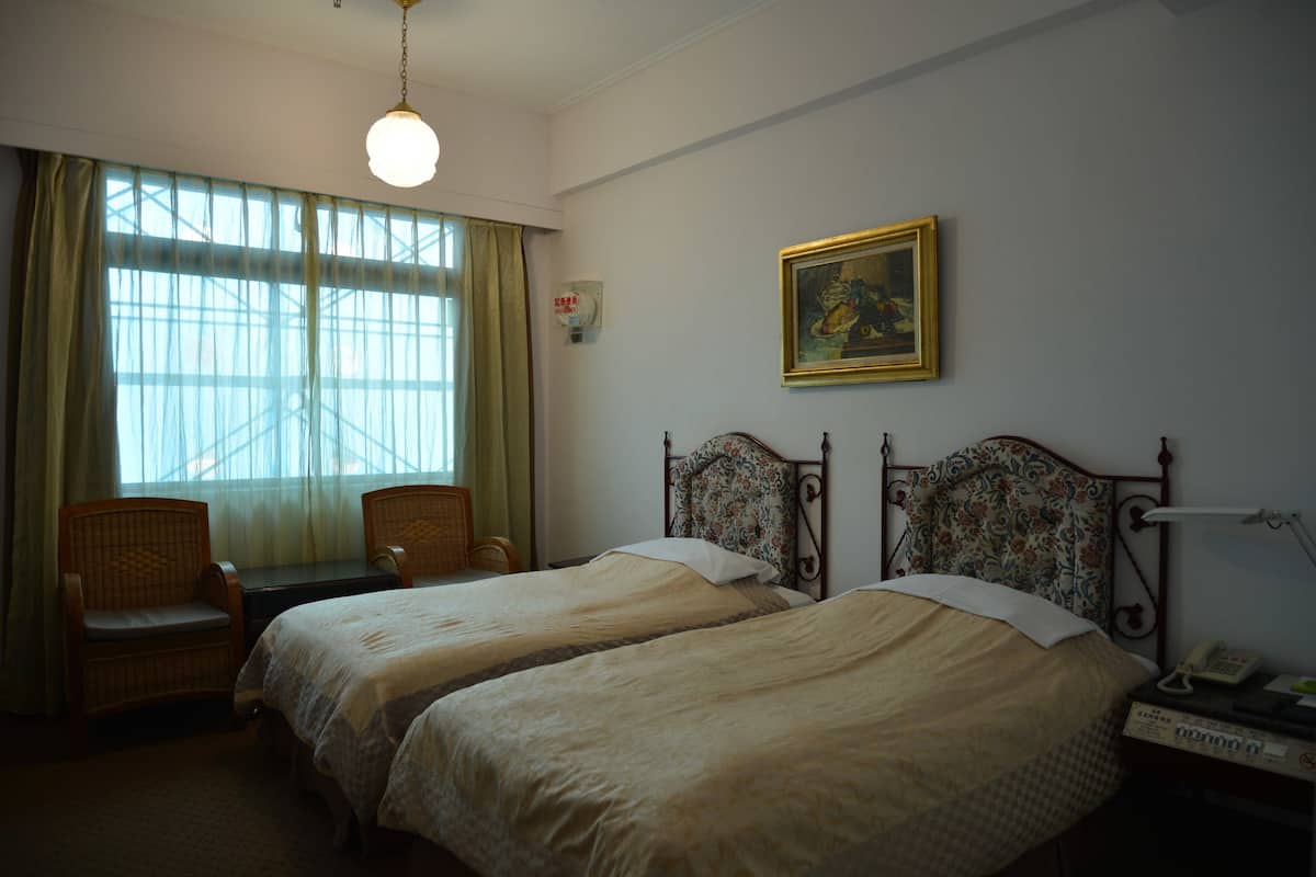 A hotel room with two beds, a desk, chairs, a painting on the wall, and a window with curtains.