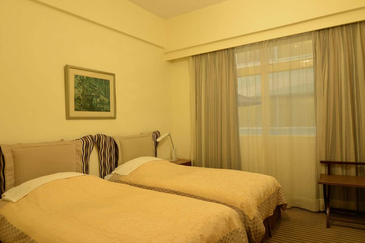 A hotel room with two beds, a desk, a chair, a lamp, a painting on the wall, and a window with curtains.